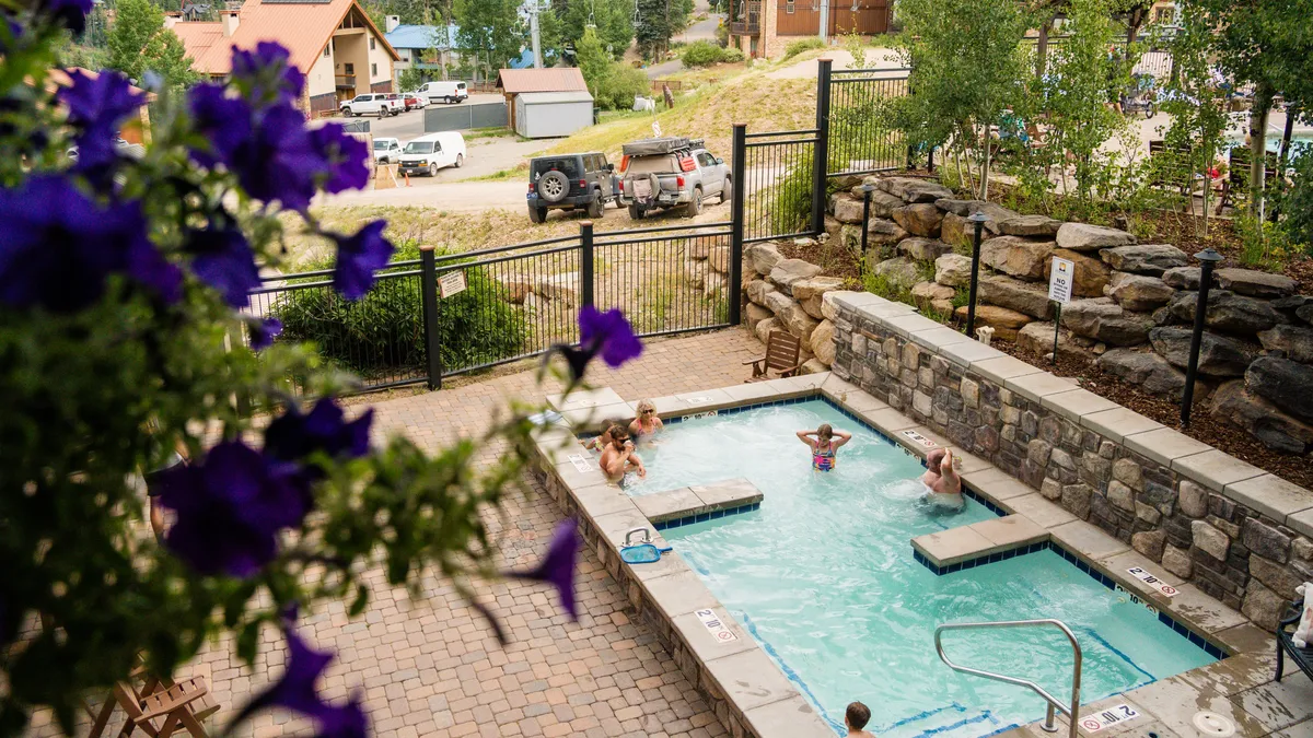 This image shows the pool area at Purgatory Lodge, featuring a sparkling swimming pool, a hot tub set amidst a stone wall, vibrant flowers, and people enjoying leisurely activities like sunbathing, swimming, and relaxing in the hot tub. The overall atmosphere is lively and cheerful, evoking the feeling of a fun resort getaway just steps from your accommodations.