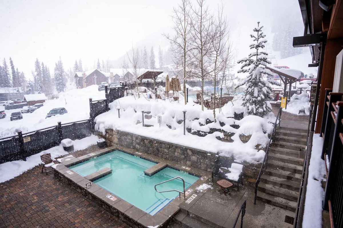 This image features an outdoor hot tub surrounded by a serene, snow-covered landscape. Pine trees and wooden structures frame the scene, and the overall mood is tranquil and inviting. The hot tub steams gently in the winter air, suggesting a cozy, restorative experience after a day on the slopes.