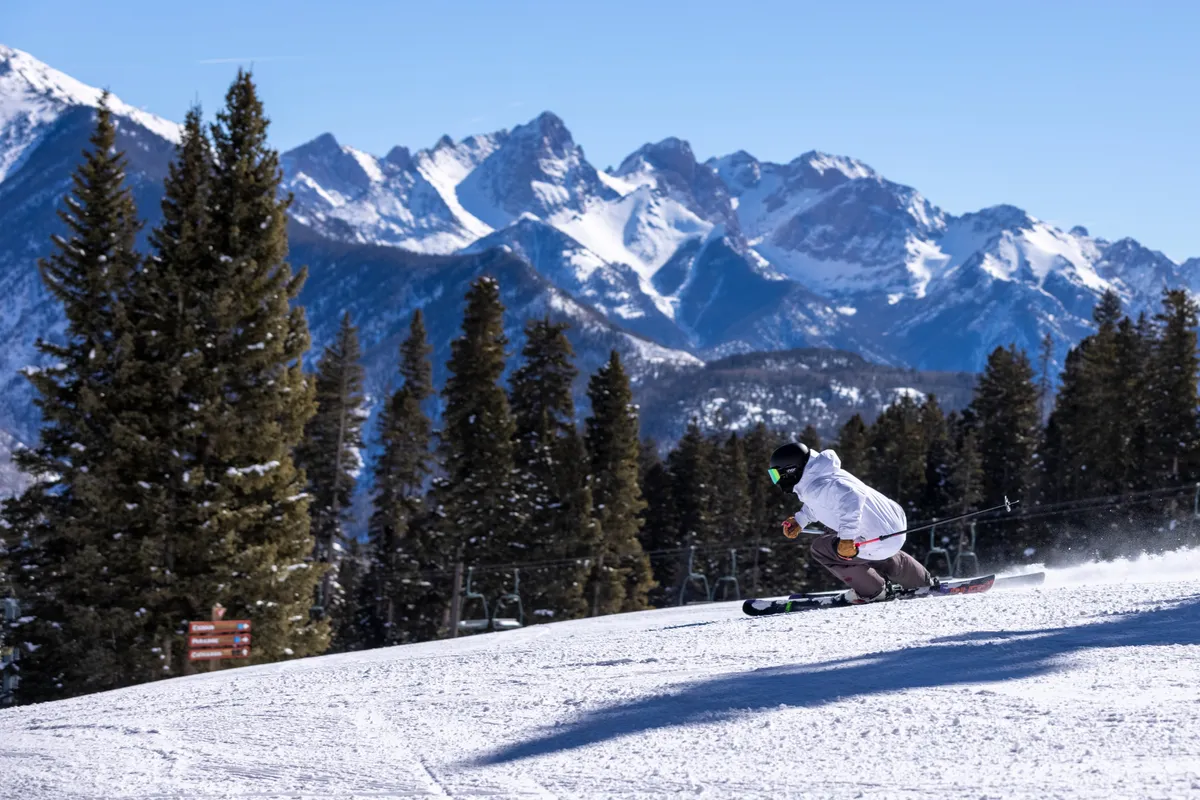 A dynamic exterior shot featuring a skier carving down a snow-covered slope with a dramatic mountain range in the background. The excitement and energy of winter sports at Purgatory Resort are front and center, capturing the action that defines family ski vacations.