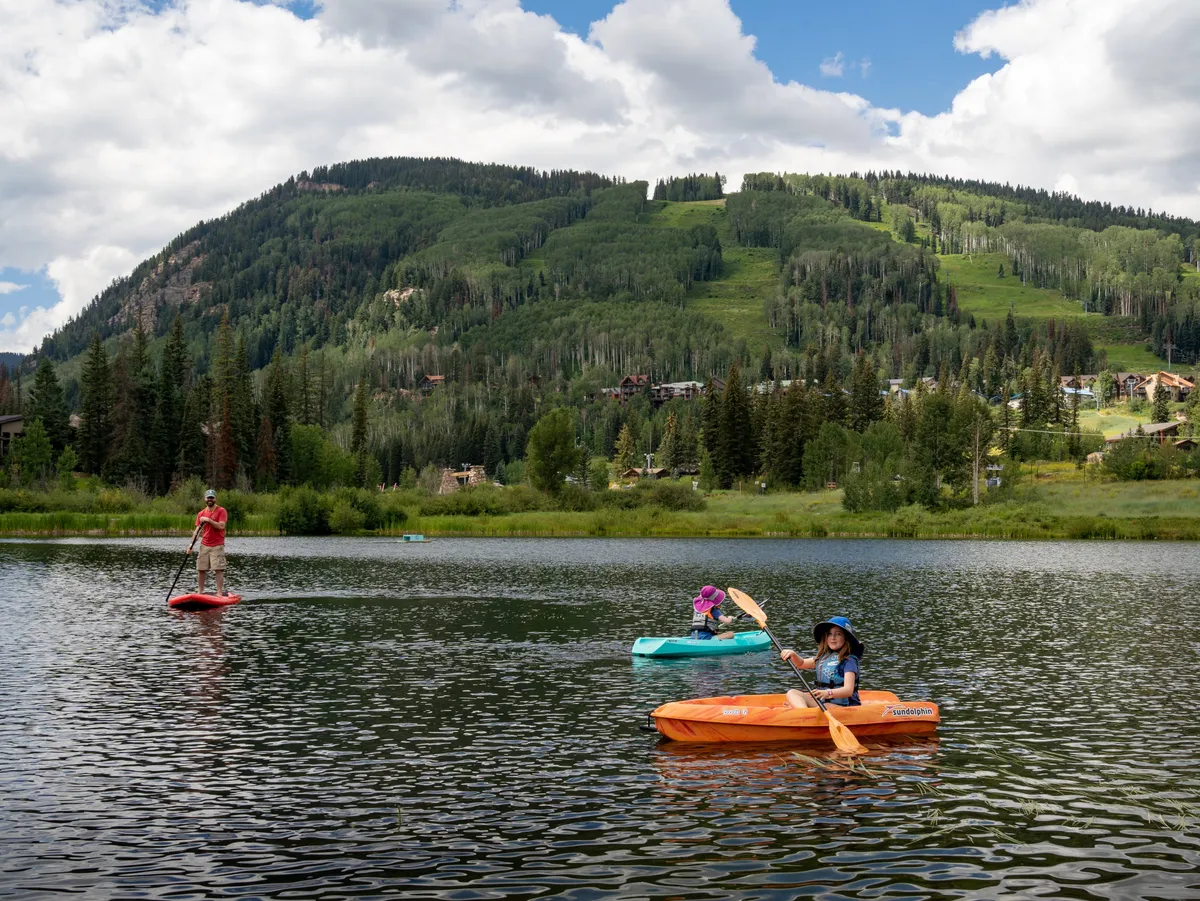 A vibrant scene of people kayaking and paddle boarding on a serene mountain lake, surrounded by lush greenery and rugged peaks. The water mirrors the stunning landscape, capturing the joyful energy of outdoor enthusiasts. This image exudes adventure and the thrill of exploring the local natural environment.