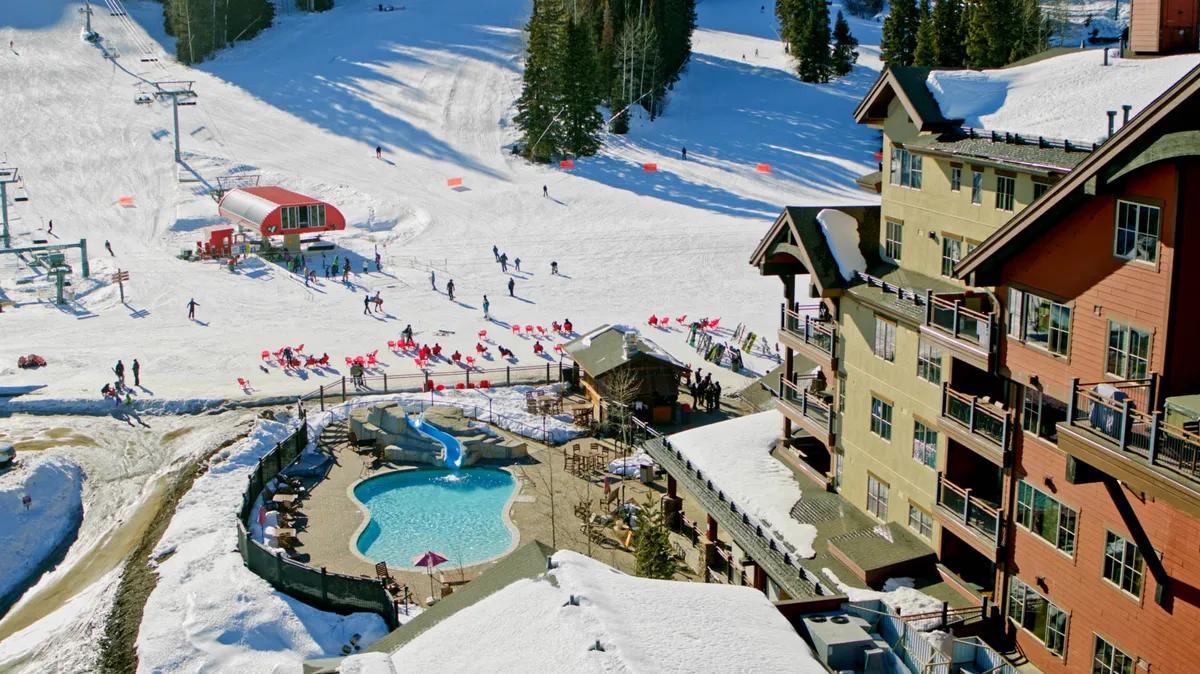 This image shows the resort building with direct sightlines to both the pool area and the adjacent ski slopes. The scene buzzes with activity, with guests swimming or lounging by the pool while others can be seen engaging in winter sports. The overall mood is cheerful and active, encapsulating the seamless blend of recreation and relaxation.