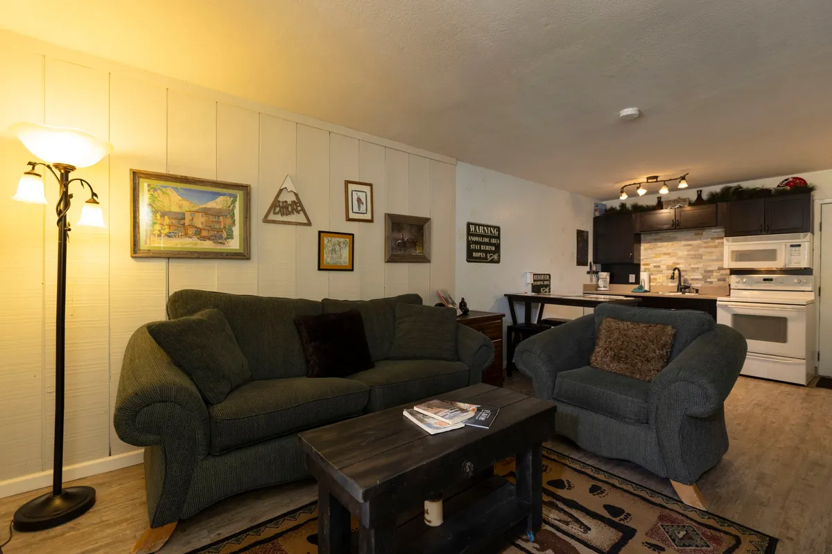 The image showcases a cozy living space with a plush sofa and inviting armchair arranged around a coffee table, complemented by decorative wall art and a view of the open kitchen area. Warm lighting creates a welcoming ambiance, perfect for relaxing, entertaining, or enjoying quiet moments after a day on the slopes.